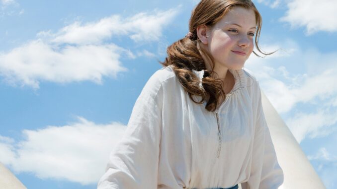 Georgie Henley is playing lucy. She is looking over the side of the Dawn Treader with a partially cloudy blue sky in the background.