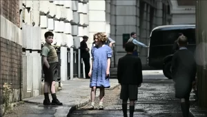 A candid, behind-the-scenes photo on a film set. Two child actors, one playing Digory and the other Polly, stand on a wet urban street. The boy wears a knickerbocker-style outfit with a sweater vest, and the girl wears a blue dress. Other cast and crew members are visible in the background.