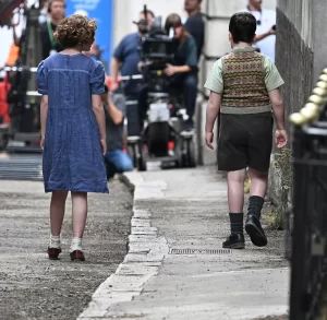 A behind-the-scenes photo of the two child actors, seen from behind, as they walk away from the camera down a narrow, wet alley on the film set. The boy is in knickerbocker-style shorts and a sweater vest, and the girl is in a blue dress with short sleeves.