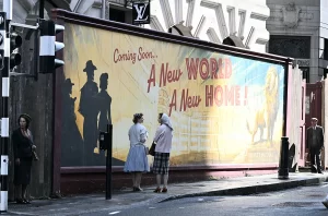 A behind-the-scenes photo of a film set showing a large billboard. The billboard reads "Coming Soon... A New World A New Home!" in a retro, 1950s style font, with a large, stylized lion on the right. Extras dressed in 1950s clothing walk on the sidewalk in front of it.